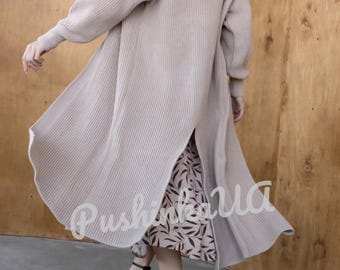 May include: A long, beige knitted cardigan with a slit up the back. The cardigan is worn over a floral print dress and the model is wearing black high heels.
