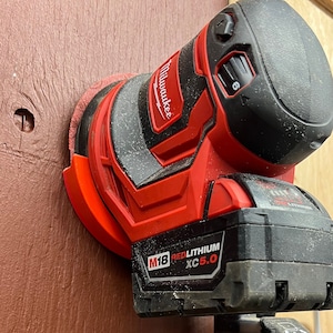Puede incluir: Una lijadora inalámbrica Milwaukee M18 roja y negra con una batería adjunta. La lijadora está montada sobre una superficie de madera de color marrón rojizo. La lijadora tiene una parte superior negra con un dial ajustado a 6.