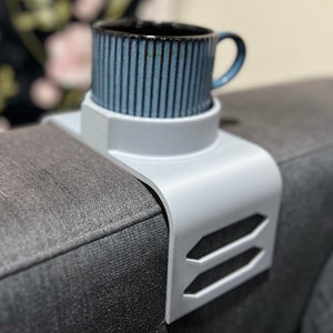Puede incluir: Una bandeja gris claro para el brazo del sofá con un portavasos incorporado, que sostiene una taza a rayas azules y negras. La bandeja tiene un diseño curvo y recortes geométricos. La taza tiene un asa azul.