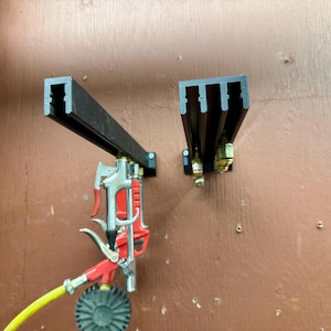 May include: Two black metal brackets mounted on a brown surface. One bracket holds a red and silver spray gun with a yellow hose and a black nozzle. The other bracket holds a brass fitting.