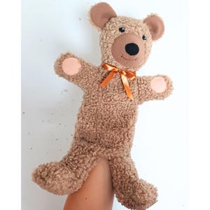 May include: A plush teddy bear puppet with a brown, textured body, brown ears, and a black nose. The bear has a light orange bow and light pink accents on its paws. The puppet is designed to be worn on the hand.