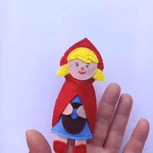 Puede incluir: Muñeca de fieltro de Caperucita Roja. La muñeca tiene una capa con capucha roja, un vestido azul y zapatos rojos. Sostiene una cesta marrón. La muñeca tiene pelo rubio y piel clara.
