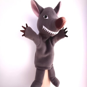 May include: A grey puppet with a toothy grin, pink ears, and brown claws. The puppet is made of soft fabric and is designed for hand use.