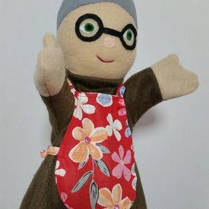 Op de afbeelding: Een handpop van een vrouw met grijs haar, die een ronde bril en een rode bloemen schort draagt. De pop is gemaakt van vilt en heeft een bruine jurk.