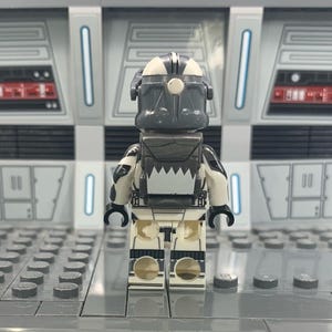 Puede incluir: Una minifigura Lego Star Wars de un clon de soldado gris y blanco con un casco y una mochila. La minifigura est&aacute; de pie sobre una superficie gris.