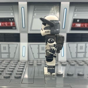 Puede incluir: Una minifigura Lego de un clon de soldado de Star Wars de color blanco y gris con una mochila negra y gris. La figura est&aacute; de pie sobre una placa base Lego gris.