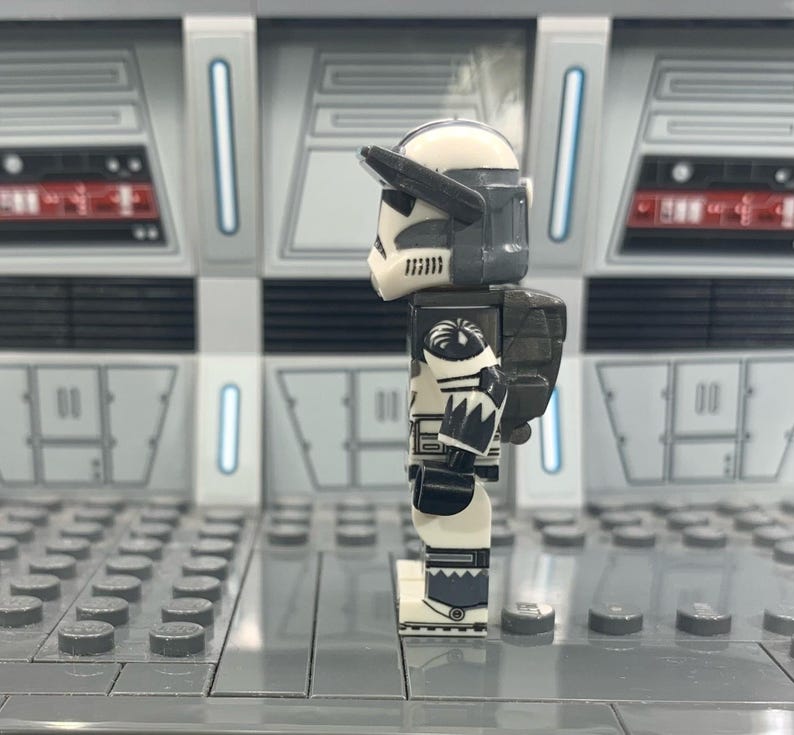 Puede incluir: Una minifigura Lego de un clon de soldado de Star Wars de color blanco y gris con una mochila negra y gris. La figura est&aacute; de pie sobre una placa base Lego gris.