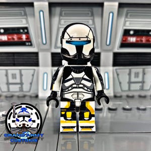 Puede incluir: Una minifigura LEGO personalizada de un soldado clon con un casco blanco y negro, una armadura blanca y negra y detalles amarillos. La minifigura está de pie sobre una superficie gris.