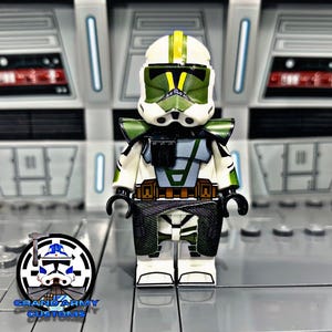 May include: A custom LEGO minifigure of a Star Wars clone trooper. The figure is wearing a white helmet with green and yellow markings, green and grey armour, and a black and green cape.