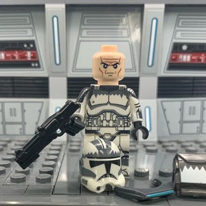 Puede incluir: Una minifigura Lego de un clon de soldado de Star Wars en blanco y negro con un rifle bl&aacute;ster negro, un casco blanco con marcas negras y una pieza de pecho blanca y negra. La minifigura est&aacute; de pie sobre una placa base Lego gris.