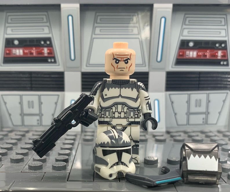 Puede incluir: Una minifigura Lego de un clon de soldado de Star Wars en blanco y negro con un rifle bl&aacute;ster negro, un casco blanco con marcas negras y una pieza de pecho blanca y negra. La minifigura est&aacute; de pie sobre una placa base Lego gris.