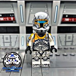 Peut inclure: Une figurine Lego de clone trooper Star Wars blanche et jaune avec un casque usé et une pièce de poitrine jaune. La figurine est debout sur une surface grise.