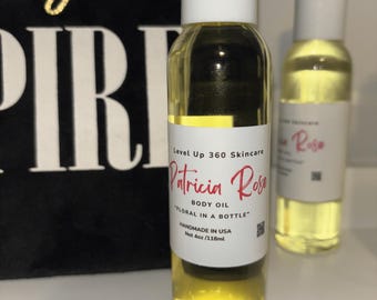 May include: Two clear glass bottles of body oil with white caps and labels. The labels read "Patricia Rose BODY OIL FLORAL IN A BOTTLE" and "HANDMADE IN USA Net 4oz /118ml". A black bag with gold text is in the background.