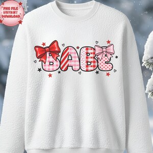 Könnte beinhalten: Weißes Sweatshirt mit dem Wort "BABE" in verspielter Schrift. Die Buchstaben sind mit Schleifen, Herzen und Sternen in Rot und Rosa verziert. Das Sweatshirt hat eine strukturierte Oberfläche.