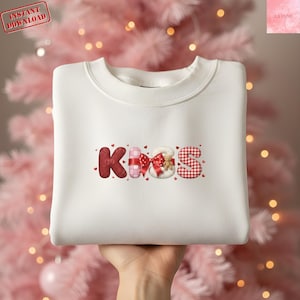 Peut inclure: Un sweat-shirt blanc avec le mot "KISS" dans une police décorative. Les lettres sont rouges, roses et blanches, avec des cœurs et des nœuds. Le sweat-shirt a un col rond et des manches longues. Le texte "Instant Download" est dans un encadré rouge en haut à gauche.