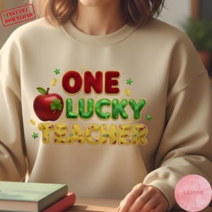 Peut inclure: Sweat-shirt beige avec l'inscription "ONE LUCKY TEACHER" en rouge, vert et jaune. Le motif comprend une pomme rouge avec un trèfle à quatre feuilles et des pièces d'or. Le texte "Instant Download" est en haut à gauche.