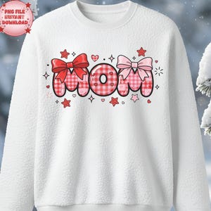 Peut inclure: Sweat-shirt blanc avec le mot "MOM" dans un motif à carreaux rose et rouge. Les lettres sont décorées de nœuds, de cœurs et d'étoiles. Le sweat-shirt a un aspect texturé et flou.