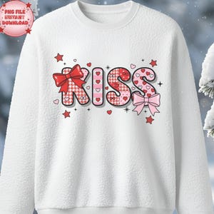 Peut inclure: Sweat-shirt blanc avec le mot "KISS" dans une police décorative. Les lettres sont ornées de cœurs, de nœuds et d'étoiles. Les lettres "K" et "S" sont à motifs, et le sweat-shirt a un aspect texturé.
