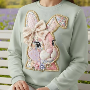 Peut inclure: Un sweat-shirt vert clair orné d'un appliqué de visage de lapin décoratif. Le lapin est fait de tissus à motifs floraux et à pois, avec un nœud et des perles. Le design comprend des couleurs rose, lavande et blanc, parfait pour Pâques ou le printemps.