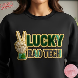 Peut inclure: Sweat-shirt noir avec l'inscription "LUCKY RAD TECH". Le motif comprend un geste de la main en signe de paix avec un trèfle, et le mot "LUCKY" en vert et or. Les mots "RAD TECH" sont en noir et or.