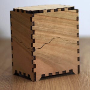 May include: A wooden box with a mountain range design etched on the front. The box is constructed with interlocking joints and has a natural wood finish. The box is sitting on a wooden surface.