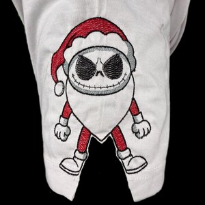 Puede incluir: Tela blanca con un diseño bordado de un personaje con gorro de Papá Noel. El personaje tiene una cara de calavera con ojos negros y una sonrisa cosida. El gorro de Papá Noel y las extremidades son rojas.