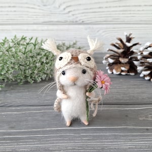 May include: A handcrafted white felt mouse wearing a tan owl hat, holding a pink flower bouquet. The mouse has long whiskers and pink feet. Decorative pine cones and greenery are in the background. The overall theme is whimsical and nature-inspired.
