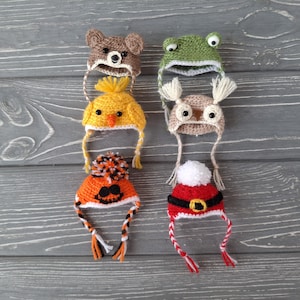 May include: A collection of six handmade crochet hats in various animal and holiday designs. The hats include a bear, frog, chick, owl, jack-o'-lantern, and Santa Claus. Each hat features unique details and earflaps with braided ties.