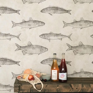 May include: A beige wallpaper featuring a repeating pattern of dark gray fish illustrations. In front, a wooden table holds a mesh bag filled with various fruits, a small bowl, and two glass bottles with labels, with the text APOSTROFO DESIGN below.
