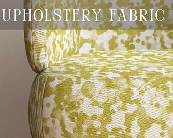 May include: A chair upholstered in a white fabric with a lime green abstract spotted pattern. The text PREMIUM UPHOLSTERY FABRIC is visible at the top, and APOSTROFO DESIGN is at the bottom. The chair features light brown wooden legs.