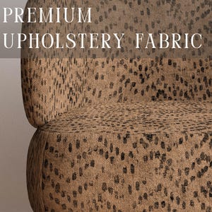 May include: A chair upholstered in a tan fabric with a black speckled animal print pattern. The text PREMIUM UPHOLSTERY FABRIC is visible at the top, and APOSTROFO DESIGN is printed at the bottom. The chair features light brown wooden legs.