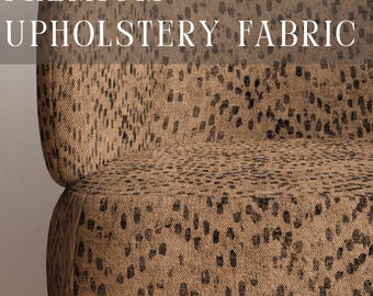 May include: A chair upholstered in a tan fabric with a black speckled animal print pattern. The text PREMIUM UPHOLSTERY FABRIC is visible at the top, and APOSTROFO DESIGN is printed at the bottom. The chair features light brown wooden legs.