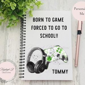 May include: A white spiral notebook with a black and green illustration of a video game controller and headphones. The text "BORN TO GAME FORCED TO GO TO SCHOOL!!" is printed in black on the front of the notebook. The name "TOMMY" is printed in black at the bottom of the notebook.