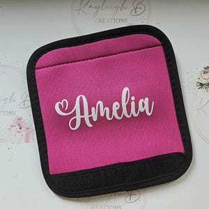 May include: Pink fabric car seat handle cover with black trim and the name Amelia in white with a heart.
