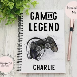 May include: White spiral-bound notebook with the words "GAMING LEGEND" in bold black letters. A graphic of a black game controller and headphones is below the text. The name "CHARLIE" is printed at the bottom. A pink pen is on the right.