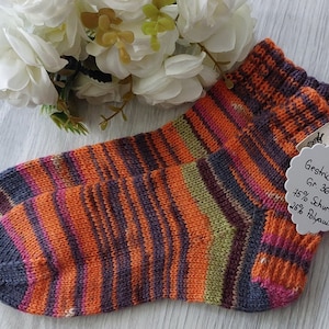 May include: A pair of hand-knitted socks in a colourful striped pattern of orange, pink, green, and purple. The socks are made with 75% wool and 25% polyamide. The tag attached to the socks reads "Gestrckte Socken Gr. 36/37 75% Schurwolle 25% Polyamid".
