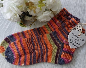 May include: A pair of hand-knitted socks in a colourful striped pattern of orange, pink, green, and purple. The socks are made with 75% wool and 25% polyamide. The tag attached to the socks reads "Gestrckte Socken Gr. 36/37 75% Schurwolle 25% Polyamid".