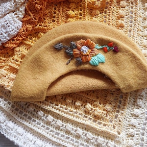 Puede incluir: Boina color mostaza con un diseño floral bordado. La flor tiene detalles en naranja, azul y blanco. La boina se muestra sobre una superficie texturizada de ganchillo con un degradado de colores amarillo y blanco. Tema: moda artesanal.