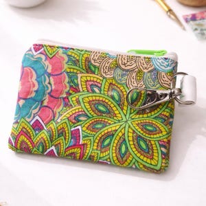 K&ouml;nnte beinhalten: Kleine, farbenfrohe Rei&szlig;verschlusstasche mit einem lebendigen Blumenmuster in Rosa, Blau, Gelb und Gr&uuml;n. Die Tasche hat einen wei&szlig;en Rei&szlig;verschluss und einen silbernen Verschluss mit einem wei&szlig;en Riemen. Dieses Accessoire ist ideal f&uuml;r kleine Gegenst&auml;nde.
