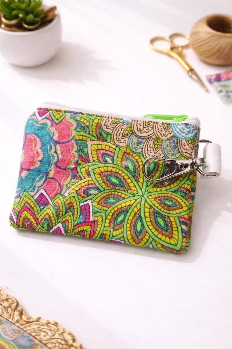 K&ouml;nnte beinhalten: Kleine, farbenfrohe Rei&szlig;verschlusstasche mit einem lebendigen Blumenmuster in Rosa, Blau, Gelb und Gr&uuml;n. Die Tasche hat einen wei&szlig;en Rei&szlig;verschluss und einen silbernen Verschluss mit einem wei&szlig;en Riemen. Dieses Accessoire ist ideal f&uuml;r kleine Gegenst&auml;nde.