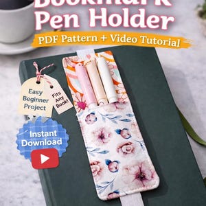 Puede incluir: Un portalápices de marcapáginas DIY con un diseño floral, que contiene tres bolígrafos. El marcapáginas está sobre un libro verde con una banda elástica blanca. Incluye el texto "DIY Bookmark Pen Holder PDF Pattern + Video Tutorial".