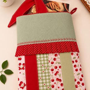 Op de afbeelding: Een rechthoekig stoffen tasje met een bovenkant van saliegroene wafelstof, een rood-witte stippenrand en een onderkant bestaande uit verticale banen in rood, groen gingham met kleine hartjes en een roze print met kersenmotieven.