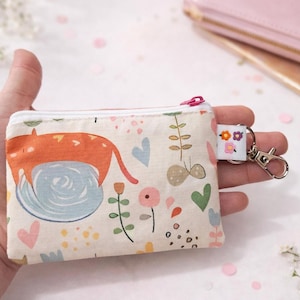 K&ouml;nnte beinhalten: Eine kleine, rechteckige Geldb&ouml;rse mit einem skurrilen Katzendesign. Die Geldb&ouml;rse hat einen cremefarbenen Hintergrund mit einem Muster aus Blumen, Herzen und Bl&auml;ttern. Sie hat einen rosa Rei&szlig;verschluss und einen kleinen Schl&uuml;sselanh&auml;nger mit einem floralen Anh&auml;nger.