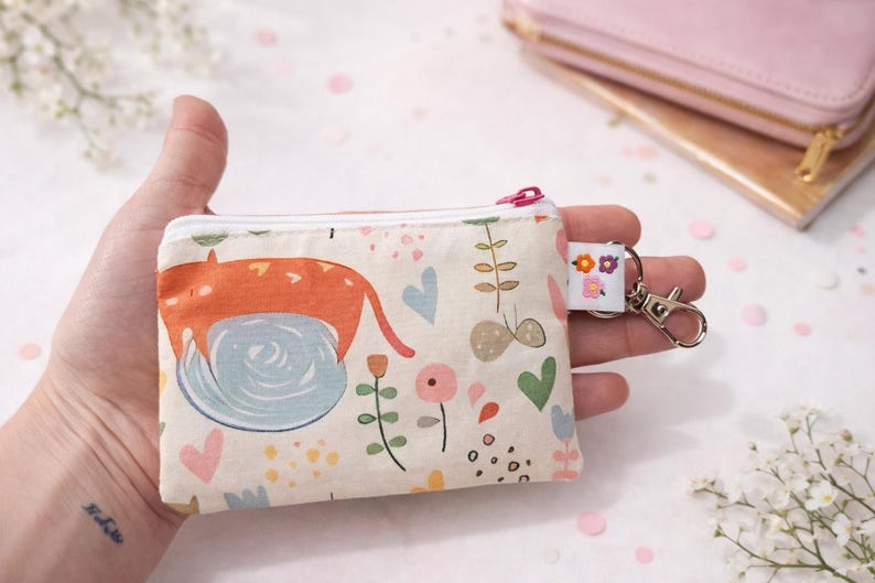 K&ouml;nnte beinhalten: Eine kleine, rechteckige Geldb&ouml;rse mit einem skurrilen Katzendesign. Die Geldb&ouml;rse hat einen cremefarbenen Hintergrund mit einem Muster aus Blumen, Herzen und Bl&auml;ttern. Sie hat einen rosa Rei&szlig;verschluss und einen kleinen Schl&uuml;sselanh&auml;nger mit einem floralen Anh&auml;nger.