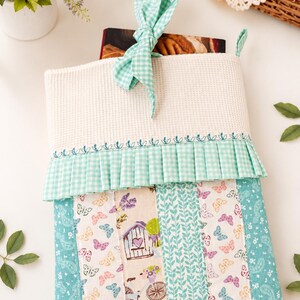 Può includere: Una custodia in tessuto per libri con una sezione superiore in tessuto a nido d'ape bianco e un nastro a quadretti verde menta. La parte inferiore è composta da pannelli verticali con stampe di farfalle, una bicicletta e motivi floreali nei toni del turchese e rosa.