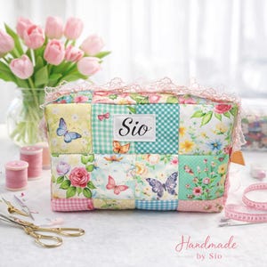 May include: A patchwork cosmetic bag with a floral and butterfly design. The bag is made from various fabrics in yellow, green, and pink, and has a lace trim. The word "Sio" is on a white label. The words "Handmade by Sio" are at the bottom.