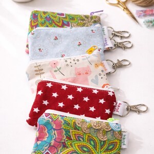K&ouml;nnte beinhalten: Eine Reihe bunter, gemusterter M&uuml;nztaschen mit silbernen Schl&uuml;sselanh&auml;ngern. Designs umfassen Blumen-, Stern- und Katzenmuster. Jede Tasche hat einen Rei&szlig;verschluss und ein kleines Etikett mit der Aufschrift "hand made".