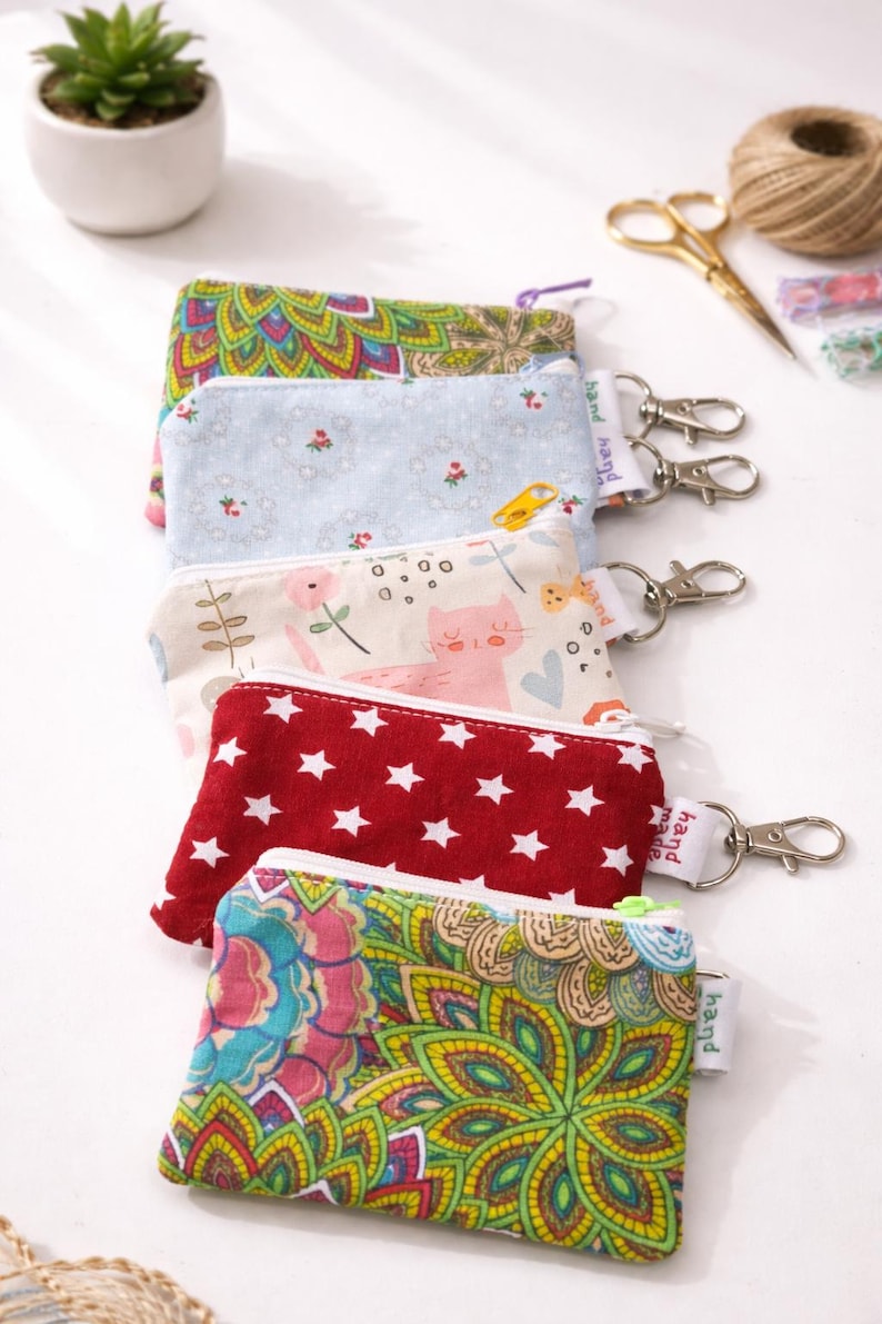 K&ouml;nnte beinhalten: Eine Reihe bunter, gemusterter M&uuml;nztaschen mit silbernen Schl&uuml;sselanh&auml;ngern. Designs umfassen Blumen-, Stern- und Katzenmuster. Jede Tasche hat einen Rei&szlig;verschluss und ein kleines Etikett mit der Aufschrift "hand made".
