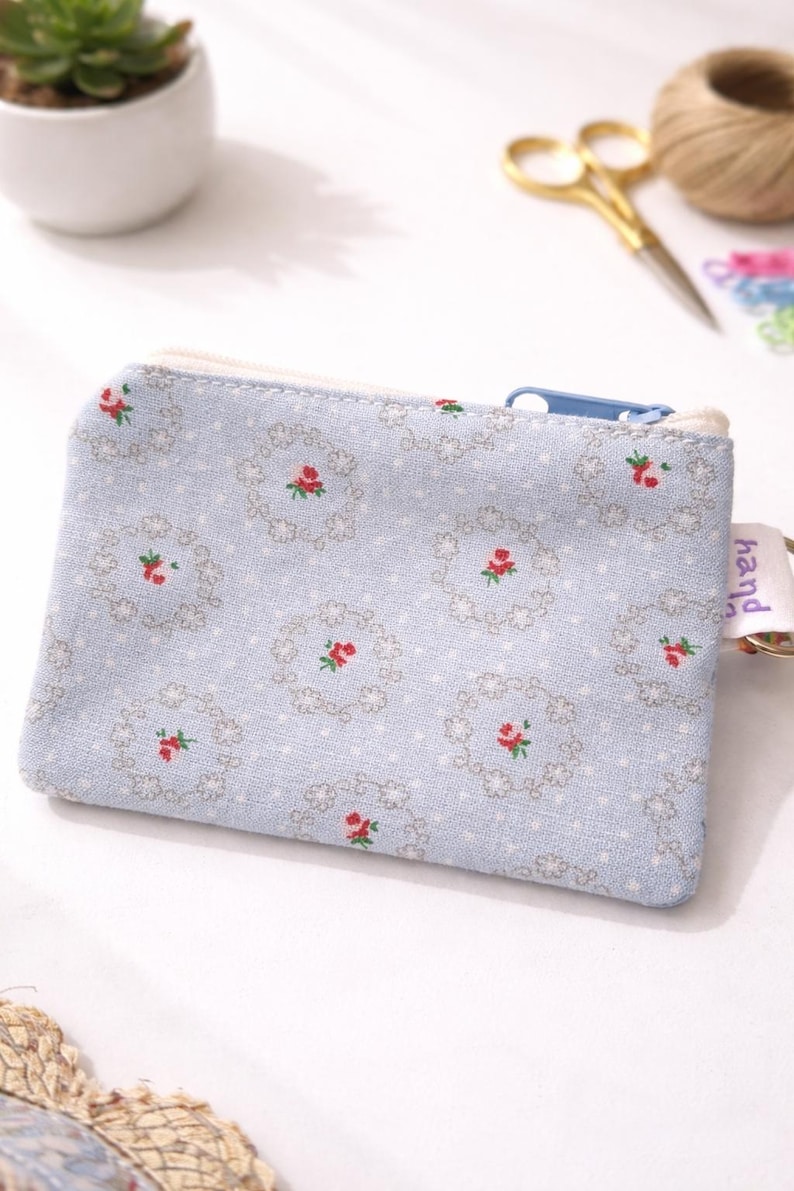 K&ouml;nnte beinhalten: Hellblaue Rei&szlig;verschlusstasche mit floralem Muster aus roten Rosen und grauen Akzenten. Die Tasche hat einen wei&szlig;en Rei&szlig;verschluss und ein kleines Etikett mit der Aufschrift "hand".