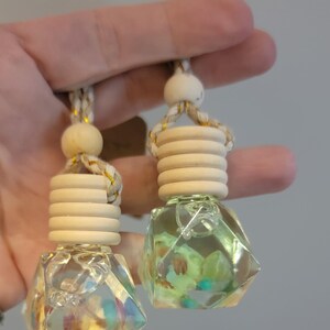 May include: Two clear, faceted glass car air fresheners with wooden tops and decorative rope hangers. The glass contains colorful, iridescent accents. The wooden tops are light-colored and cylindrical. The rope is beige with gold accents.